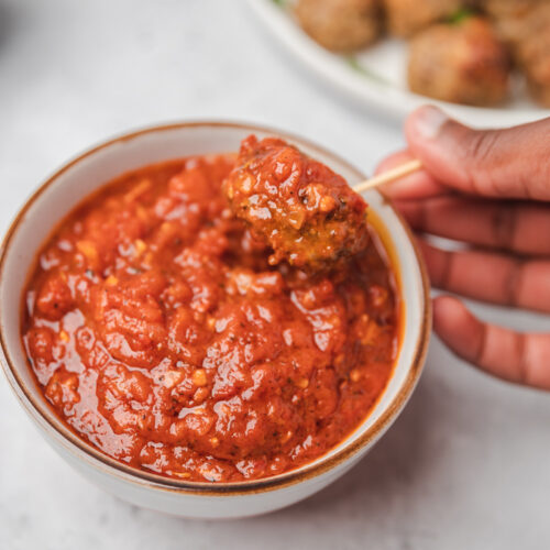 a hand dipping mini meatball into a bowl of marinara sauce, there is also a plate of meatballs placed behind it.