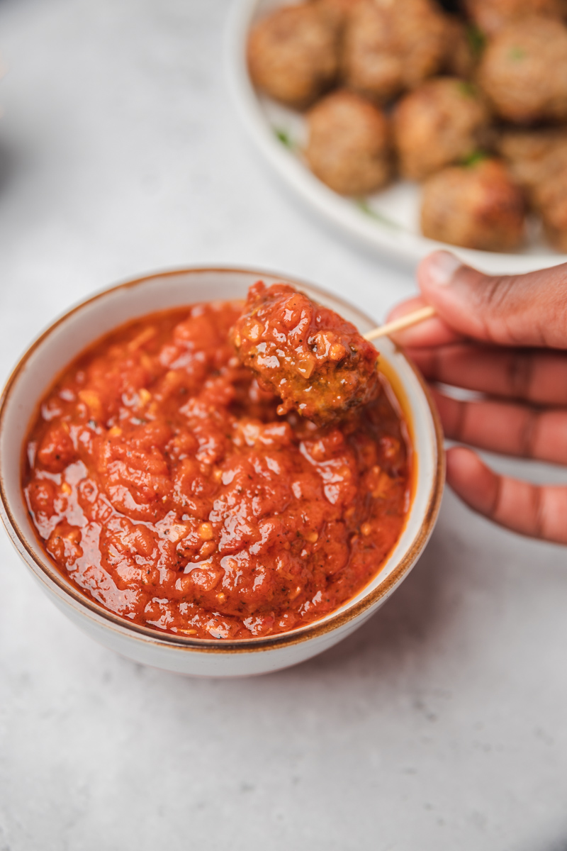 a hand dipping mini meatball into marinara sauce with a toothpick.