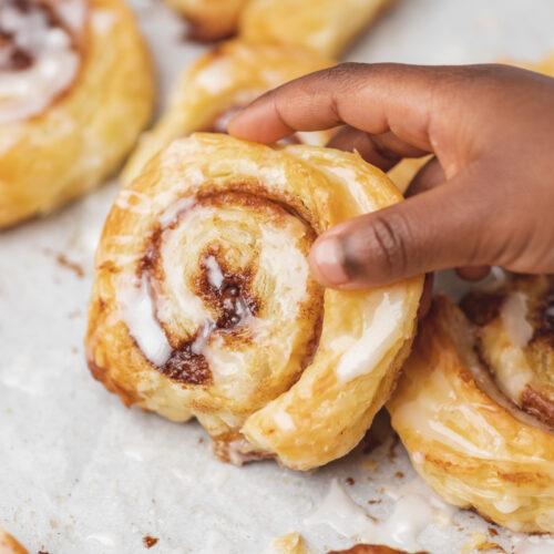 a hand holdin a glazed puf past cinnamon roll from a baking tray.