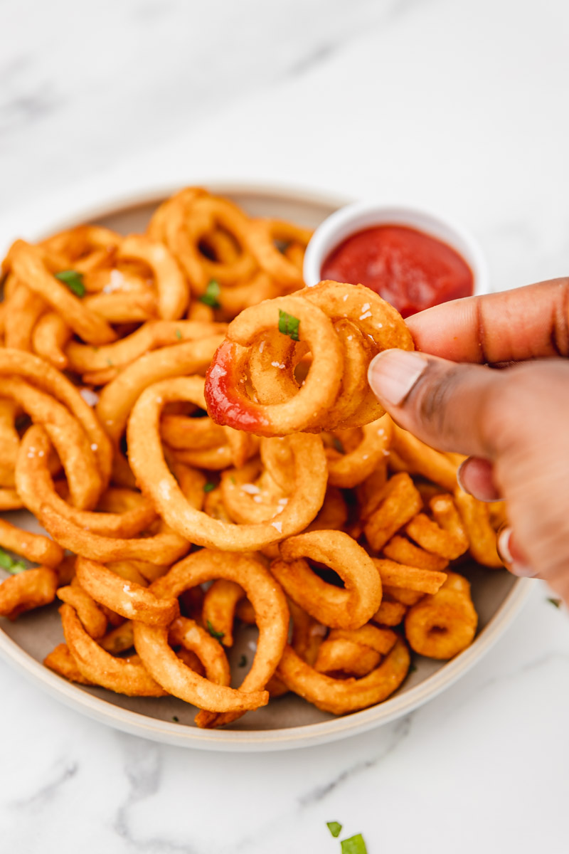 a hand holding up curly fry dipped in ketchup.