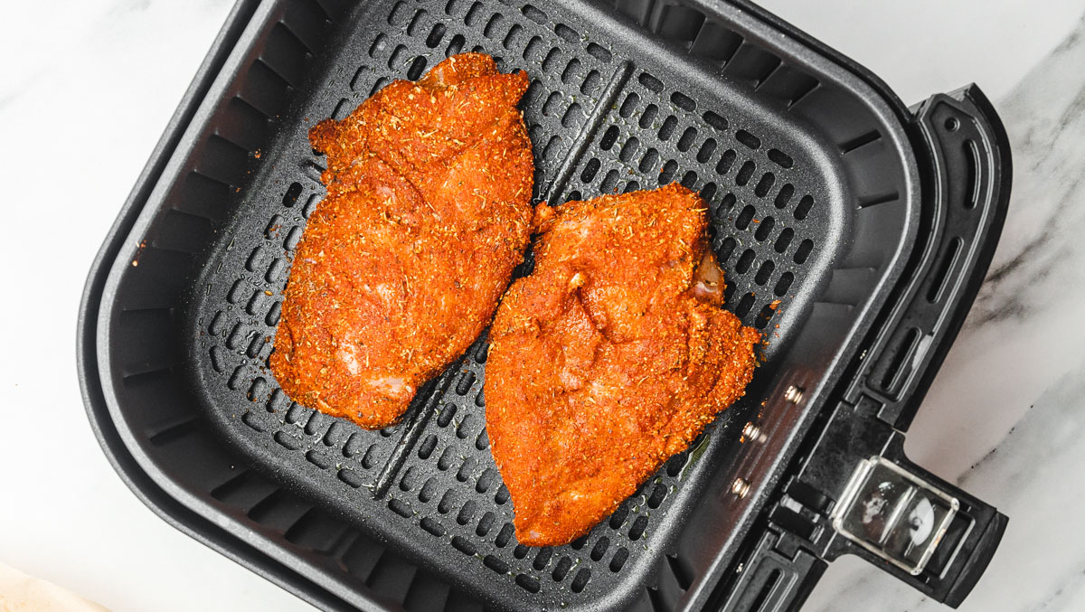 two uncooked seasoned chicken breast in air fryer basket.