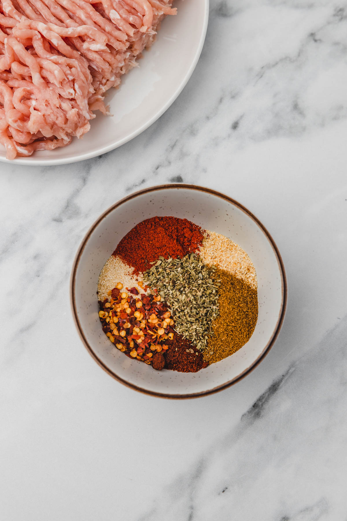 picesa and herbs in a bowl placed beside raw ground chicken.