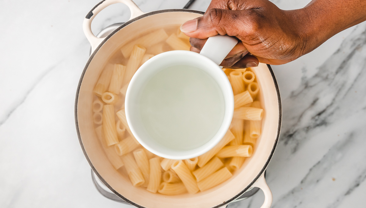a hand holding a cup of pasta water over a pot of pasta.