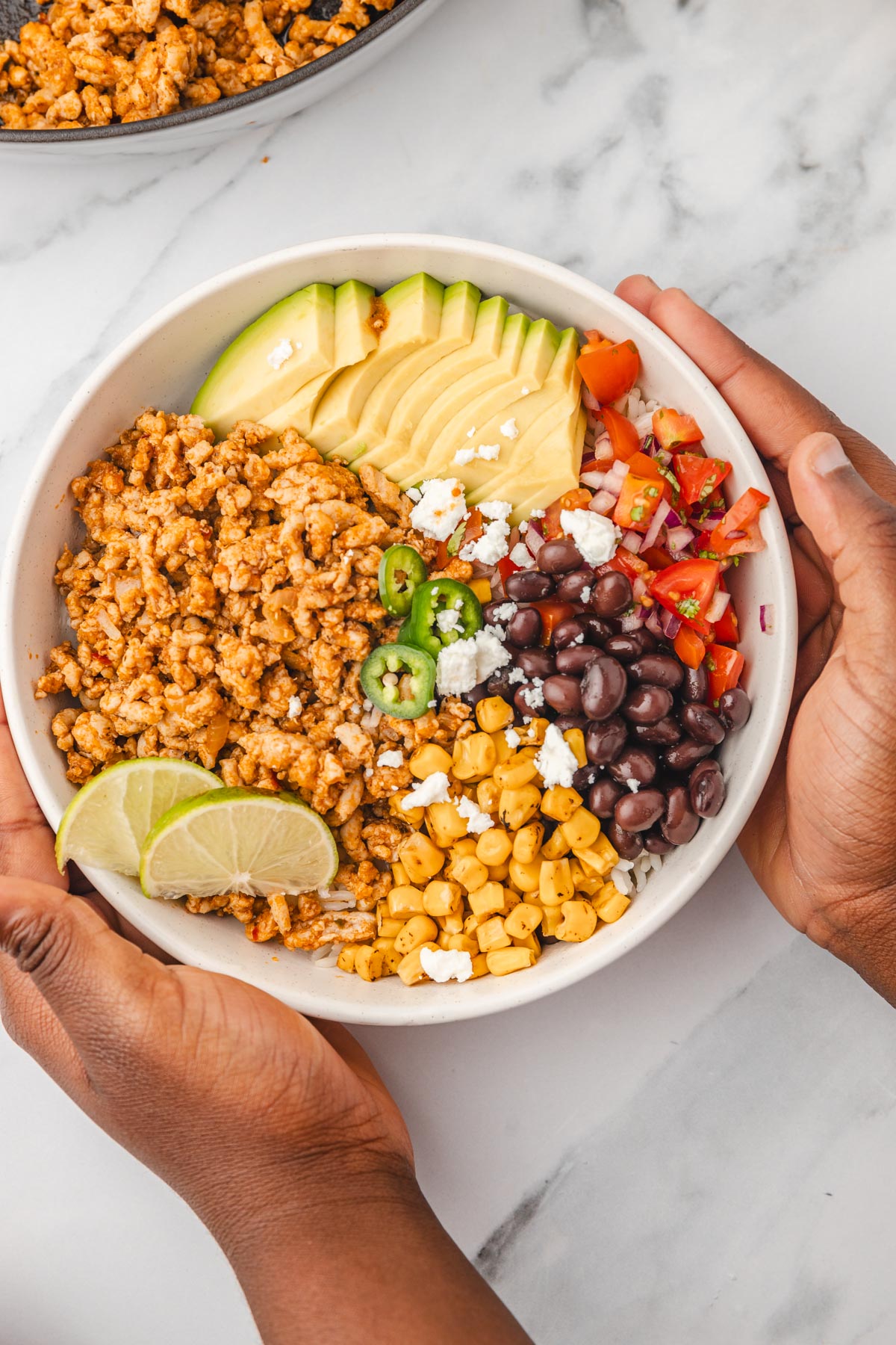 a hand lifting ground chicken rice bowl topped with avocado, charred corn, lime wedges, black beans and pico de gallo from the table.