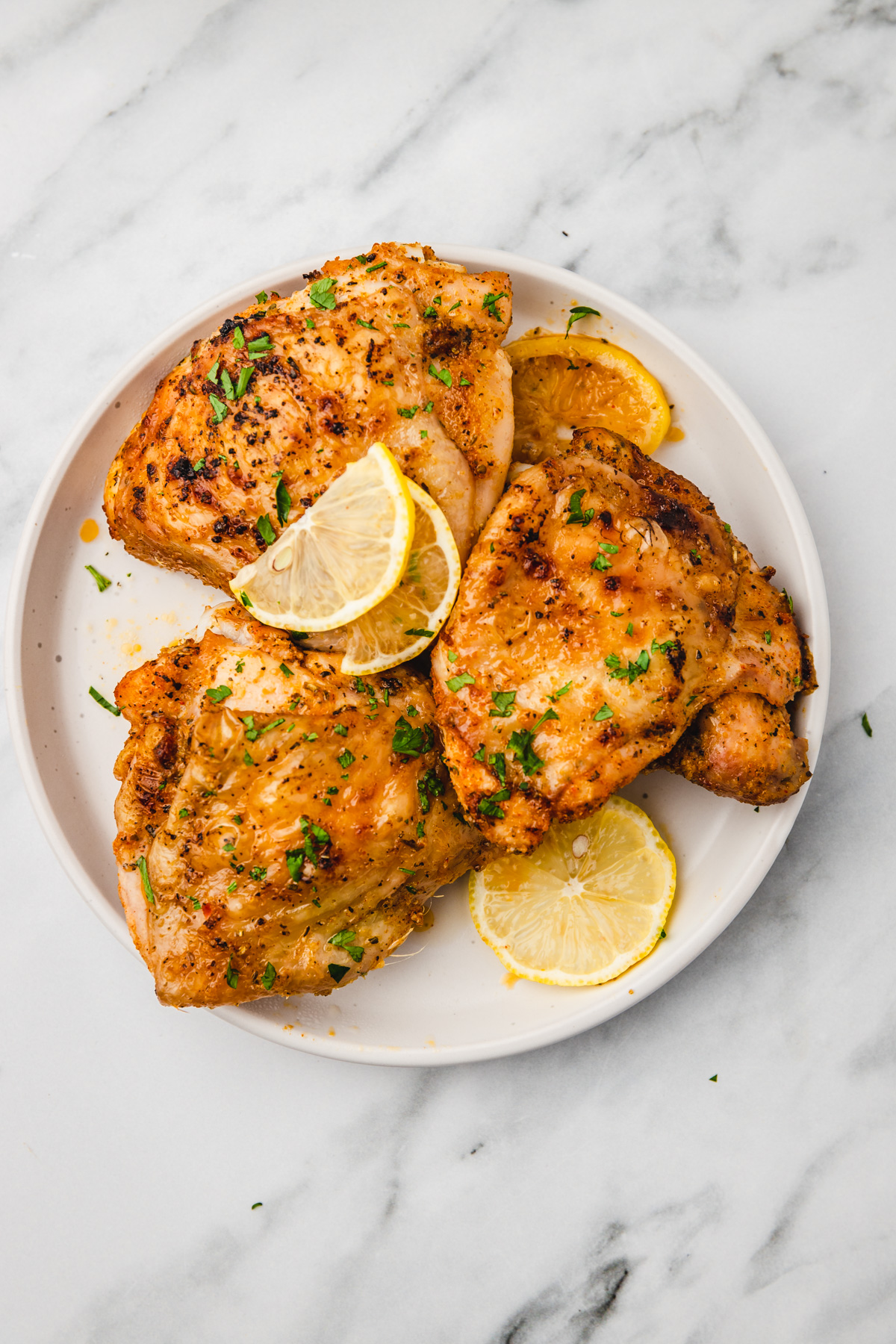three chicken thighs on a plate with lemon slices and chopped parsley.